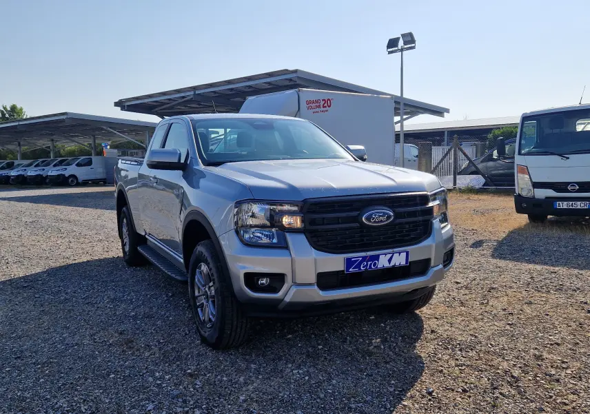 Ford Ranger Super Cabine gris iconic vu en 3/4 avant droit sur un terrain gravillonné avec plusieurs véhicules utilitaires blancs en arrière-plan.