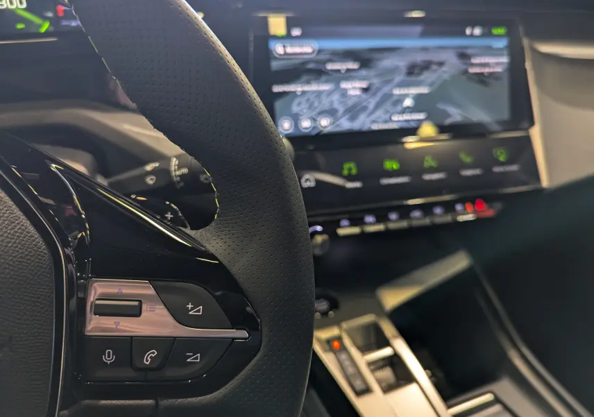 Gros plan sur le volant en cuir noir perforé de la Peugeot 308 2025 avec écran tactile et console centrale moderne en arrière-plan.