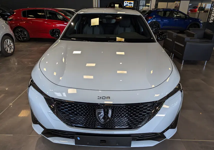 Vue frontale d'une Peugeot 308 GT blanche Okénite avec calandre noire et logo lion au centre, en showroom.