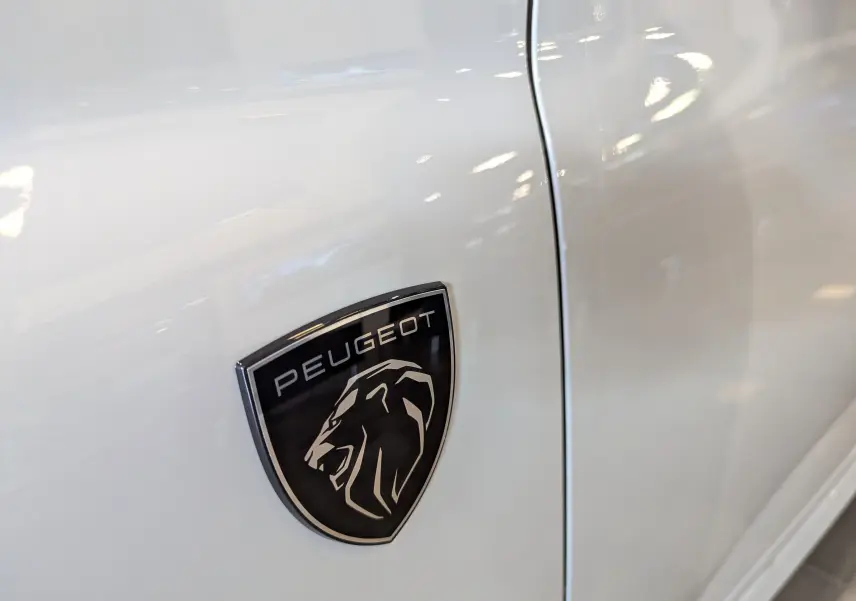 Gros plan sur l'écusson noir Peugeot avec lion argenté sur la porte blanche Okénite d'une 308 GT 2025.