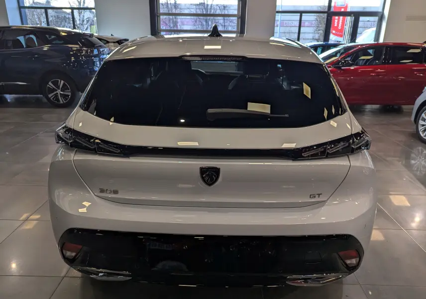 Vue arrière d'une Peugeot 308 GT blanche Okénite avec feux arrière noirs et logo lion central en concession.
