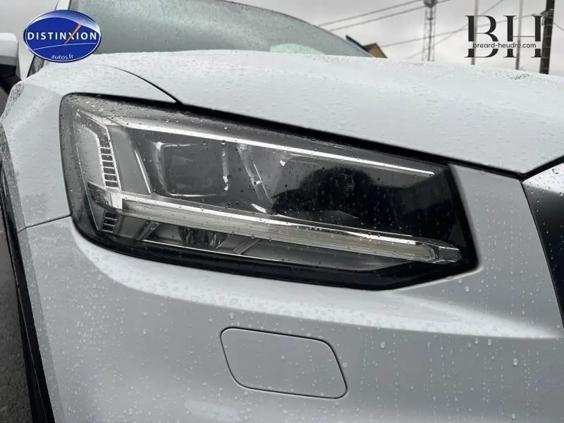 Gros plan sur le phare avant gauche blanc Ibis d'un Audi Q2 2018 sous la pluie avec détails LED visibles.