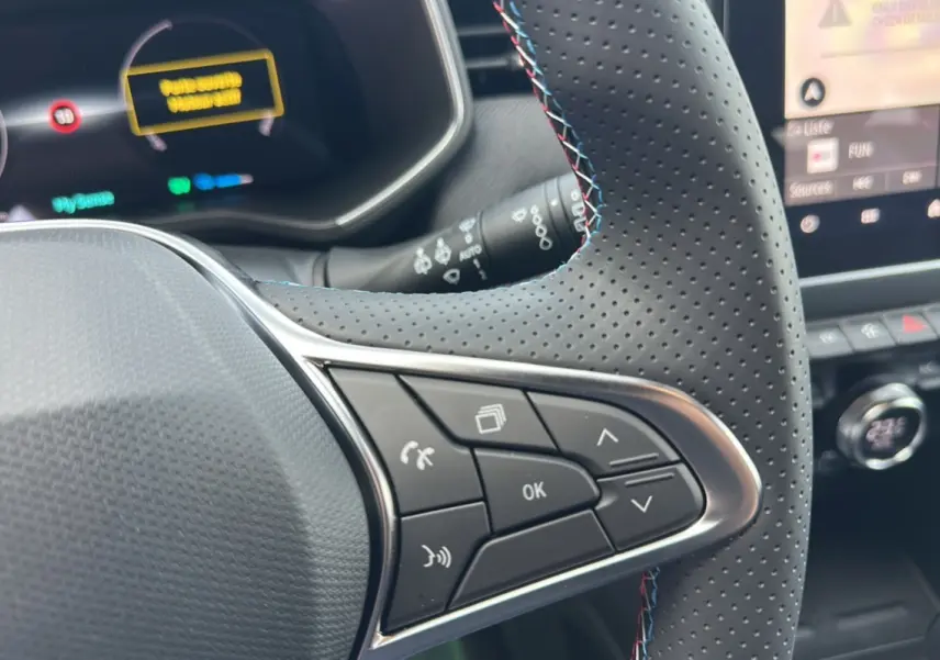 Gros plan sur les commandes multifonctions du volant cuir perforé de la Renault Clio V E-Tech gris rafale, tableau de bord en arrière-plan.