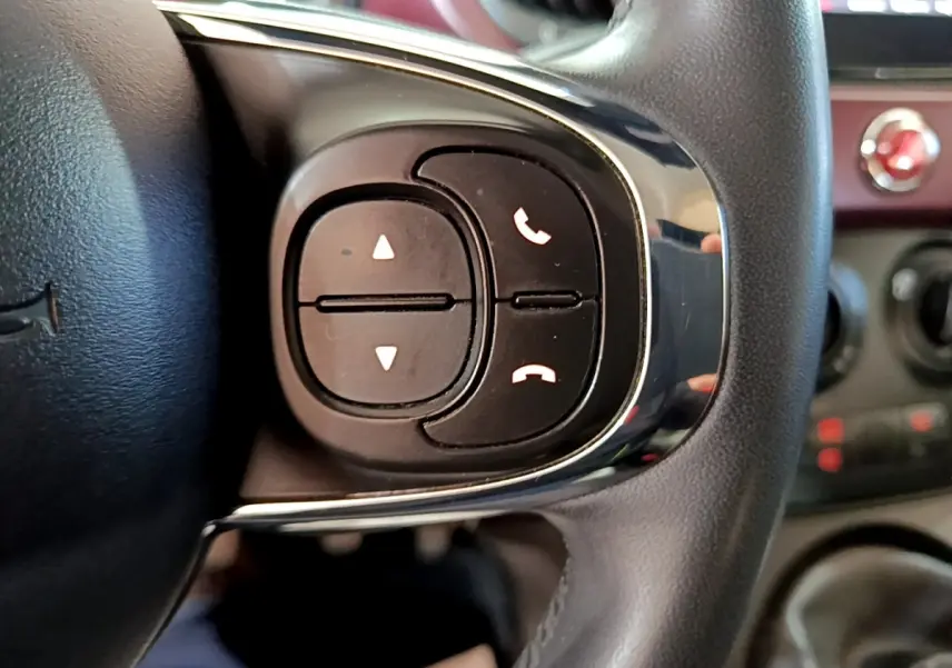 Gros plan sur les commandes noires du volant cuir de la FIAT 500 rouge, avec boutons de téléphone et réglages audio.