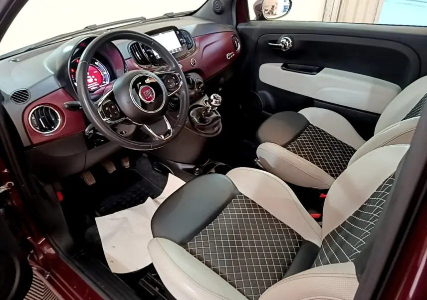 Vue intérieure côté conducteur de la Fiat 500 rouge, avec tableau de bord bordeaux et sièges bi-ton gris et noir.