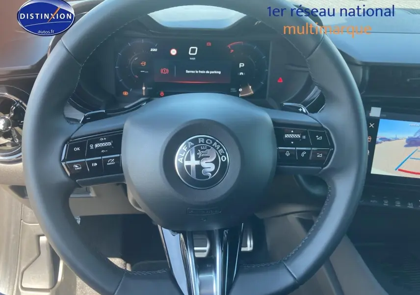 Vue rapprochée du volant noir de l'Alfa Romeo Junior 1.2 hybride avec tableau de bord numérique et écran tactile.