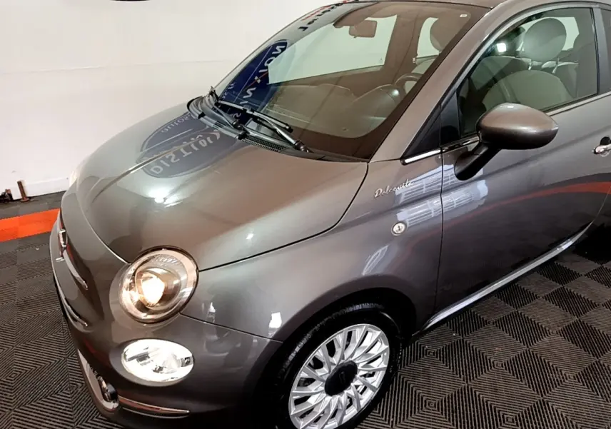 Vue 3/4 avant droit d'une FIAT 500 Dolcevita gris foncé avec phares allumés et jantes argentées détaillées.