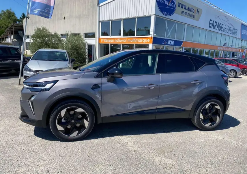 Profil côté gauche du Renault Captur gris Cassiopée avec toit noir, garé devant un garage multimarque.