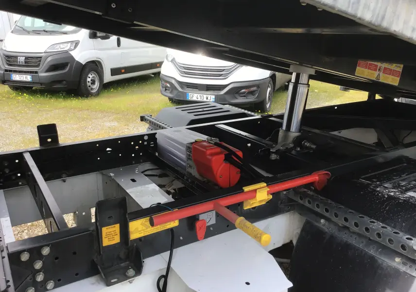 Vue rapprochée sous la benne du Renault Master blanc, montrant le châssis, vérin hydraulique et coffre de rangement.