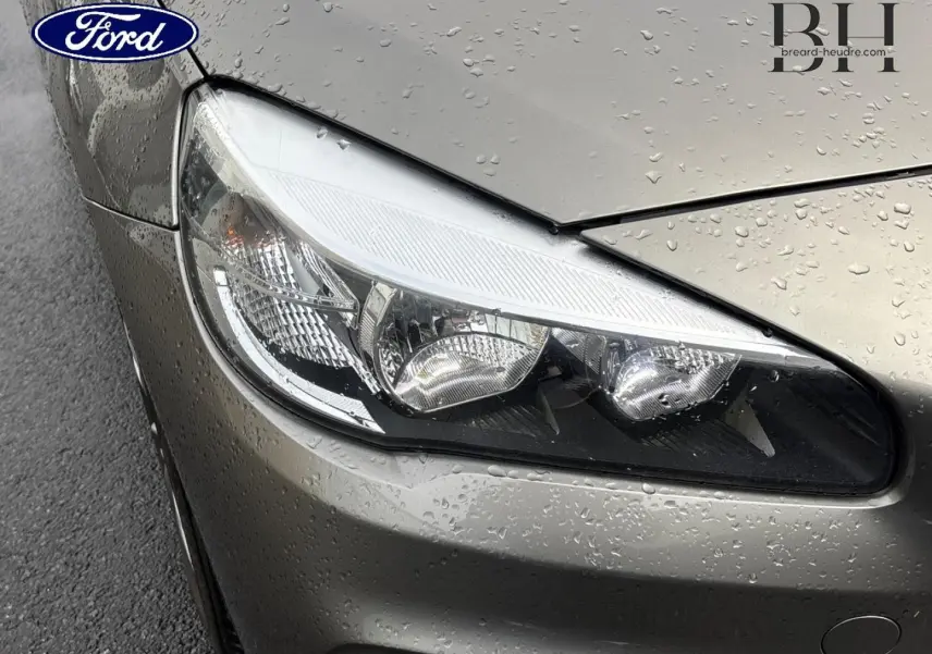 Gros plan sur le phare avant gauche d’une BMW Série 2 Active Tourer platinumsilber avec gouttes de pluie sur la carrosserie.