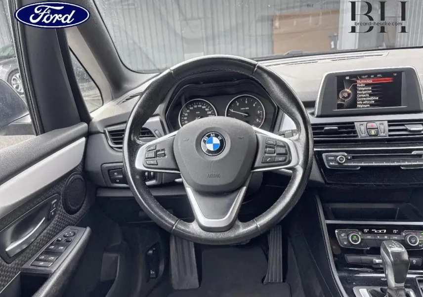 Vue intérieure centrée sur le volant cuir BMW et tableau de bord noir avec écran multifonction et commandes visibles.