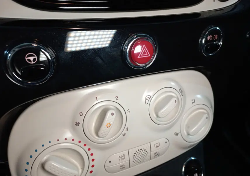 Gros plan sur les commandes de climatisation blanches et le bouton rouge de warning dans l’habitacle d’une Fiat 500 noire.