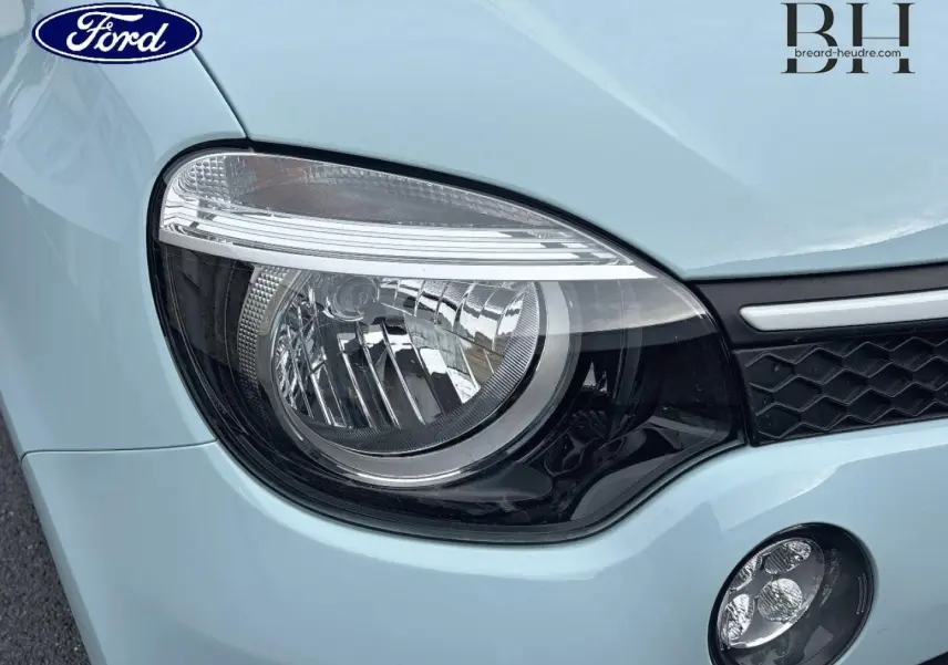 Gros plan sur le phare avant gauche d’une Renault Twingo bleu dragée avec calandre noire et logo Ford visible en reflet.