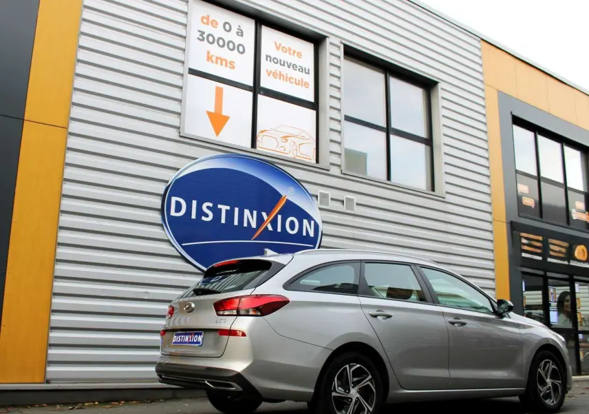 Hyundai i30 SW 2024 argent vue 3/4 arrière côté droit, stationnée devant un bâtiment commercial Distinxion.