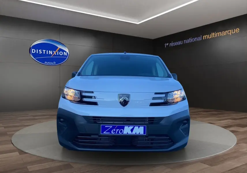 Vue frontale d'un Peugeot Partner blanc 2024 avec phares allumés et plaque "Zéro KM" en concession.