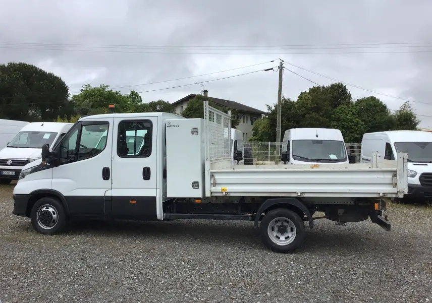 Profil côté gauche d'un Iveco Daily blanc double cabine avec benne et coffre, roues jumelées visibles sur un sol gravillonné.