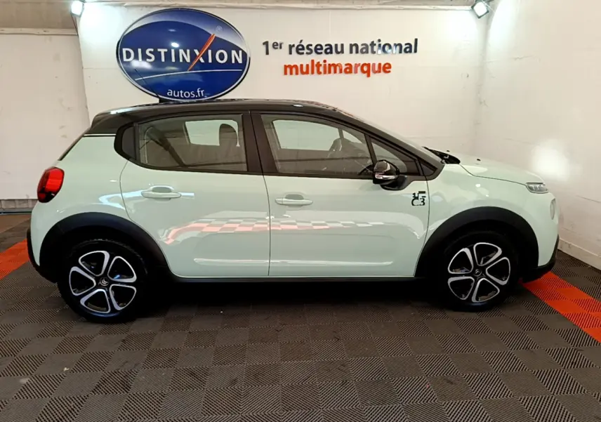 Profil côté gauche d'une Citroën C3 PureTech 82 vert clair avec toit noir et jantes stylisées en intérieur showroom.