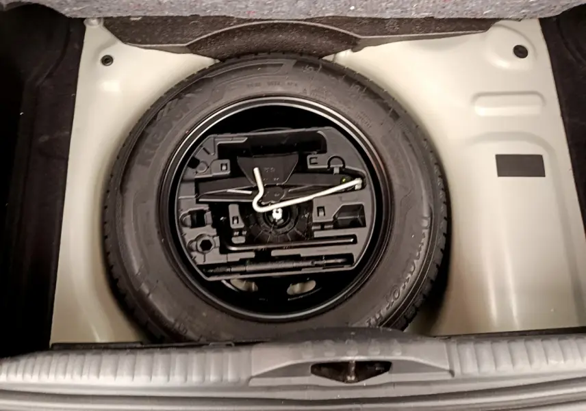 Vue plongeante sur la roue de secours noire et son kit d'outils dans le coffre beige d'une Citroën C3 verte.