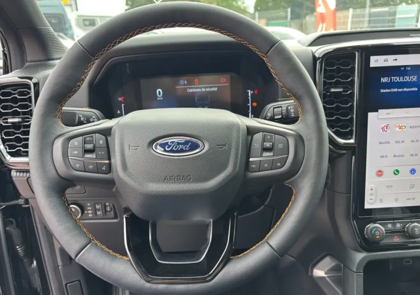 Vue rapprochée du volant noir avec surpiqûres orange du Ford Ranger 2025 Wildtrak Plus et son tableau de bord numérique.