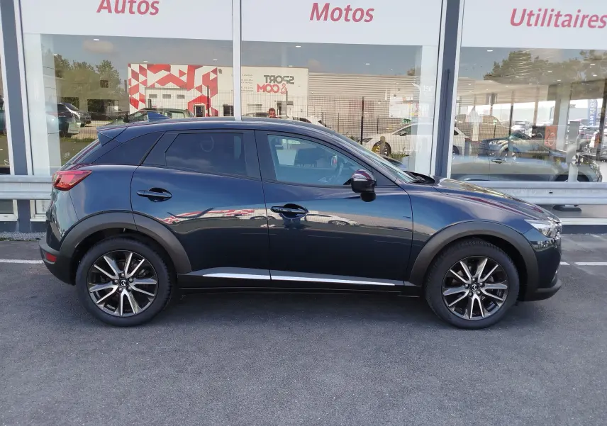 Profil côté gauche d'un Mazda CX-3 2016 bleu foncé avec jantes alliage et vitres teintées, stationné devant une vitrine.