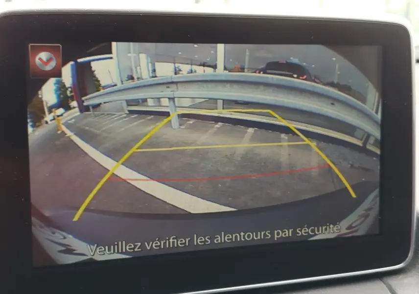 Vue caméra de recul du Mazda CX-3 bleu Dynamic Blue Mica, montrant la zone de stationnement et la barrière métallique arrière.