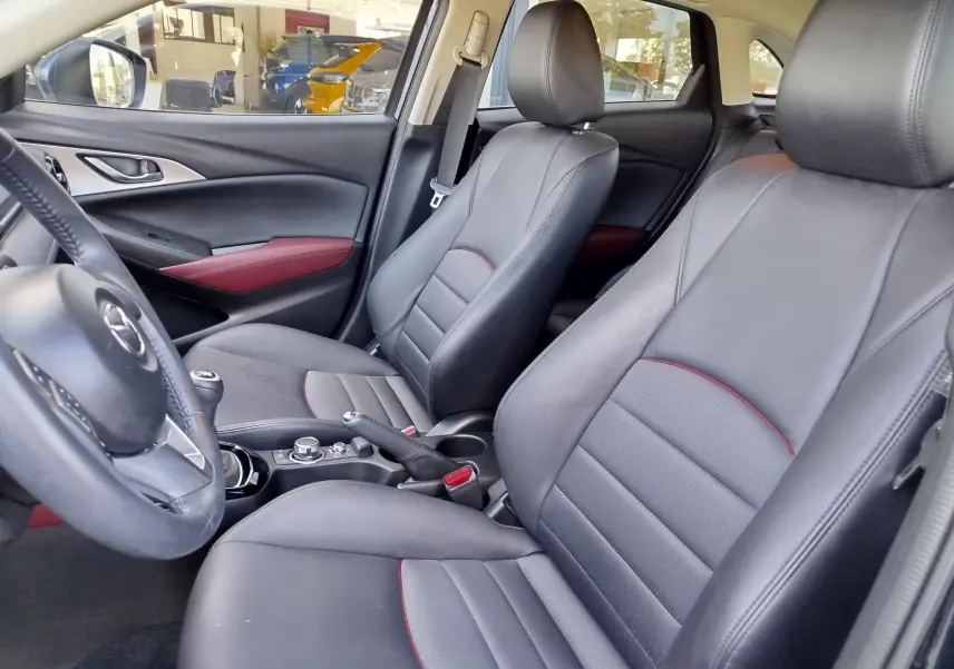 Vue intérieure côté conducteur du Mazda CX-3 2016 avec sièges cuir noir et surpiqûres rouges, volant multifonction visible.