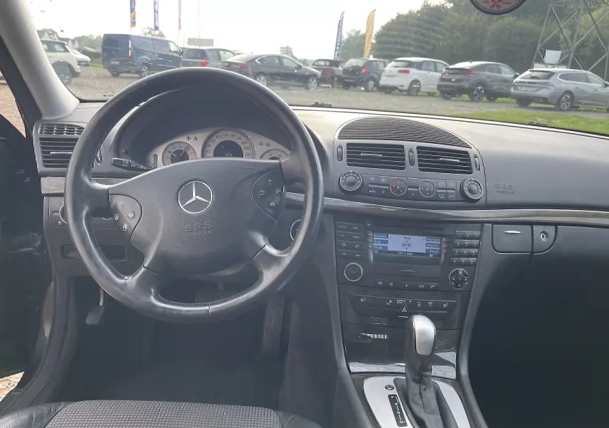 Vue intérieure centrée sur le volant et la console centrale d'une Mercedes Classe E noire de 2003 avec boîte automatique.