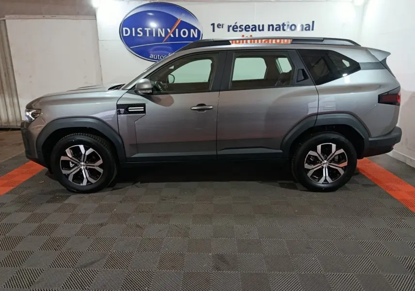 Vue de profil droit d'un SUV Dacia Bigster gris foncé 2025 avec jantes alliage et barres de toit noires.