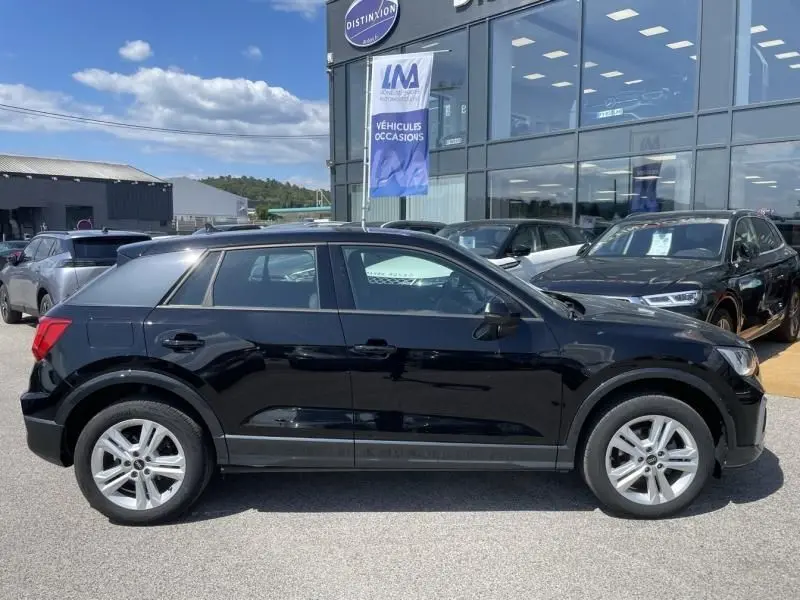 Audi Q2 TFSI 150CV noir métal vue de profil côté gauche, garé devant un concessionnaire avec d'autres véhicules.