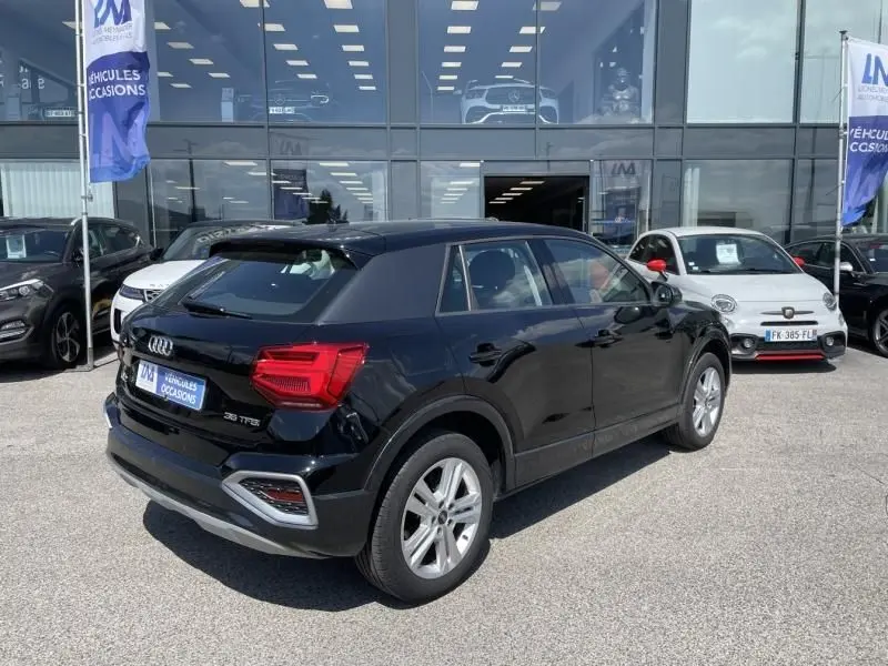 Audi Q2 noir métal vue 3/4 arrière droit, avec feux arrière rouges et jantes argentées, stationné devant un showroom.