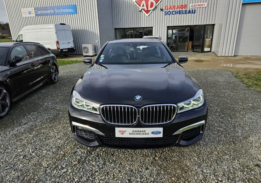 BMW Série 7 G11 noire vue de face, avec calandre double haricot et phares LED allumés devant un garage.
