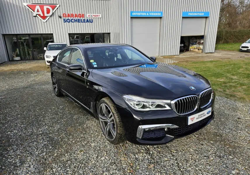 BMW Série 7 G11 noire vue en 3/4 avant droit, avec calandre chromée et jantes sport devant un garage AD Socheleau.
