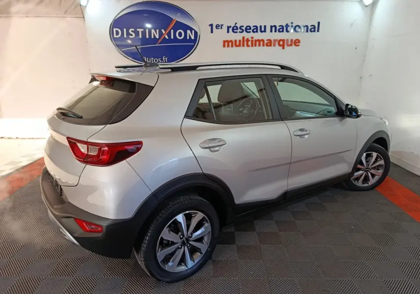 Vue 3/4 arrière droite d'un KIA STONIC gris clair 2022, avec barres de toit et jantes alliage visibles en intérieur showroom.