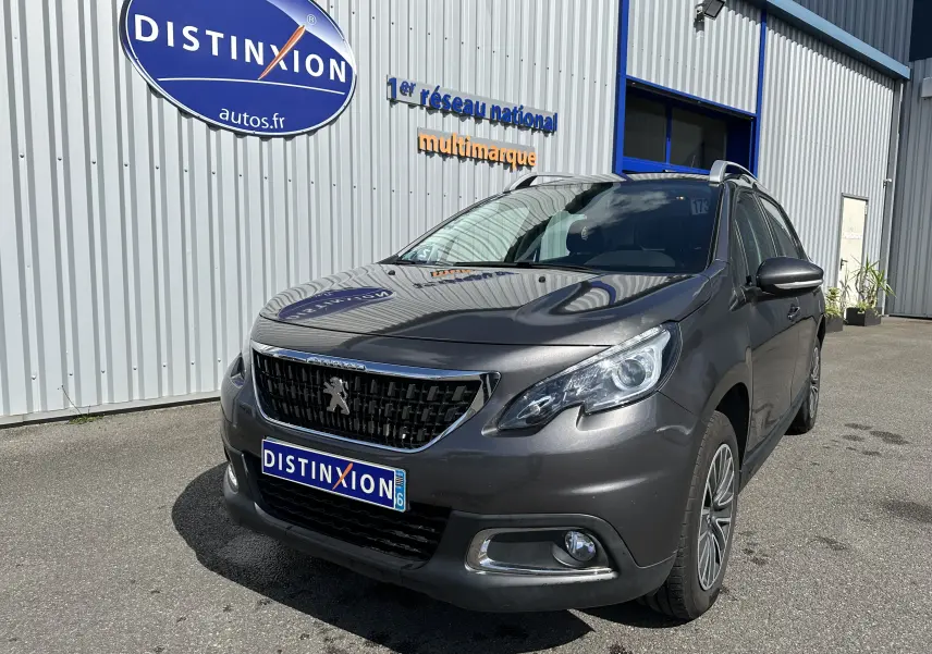 Peugeot 2008 gris 3/4 avant droit, mettant en valeur la calandre noire et les phares modernes sous un ciel ensoleillé.