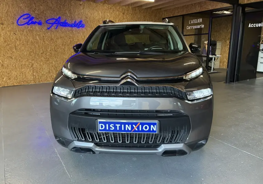 Vue frontale d'un Citroën C3 Aircross gris platinium métal avec calandre noire et plaques Distinxion en intérieur showroom.
