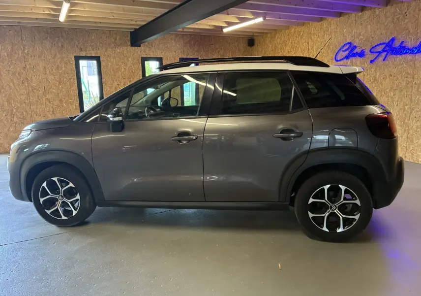 Profil côté gauche d'un Citroën C3 Aircross gris platinium métal avec toit blanc et jantes alu bi-ton en intérieur showroom.