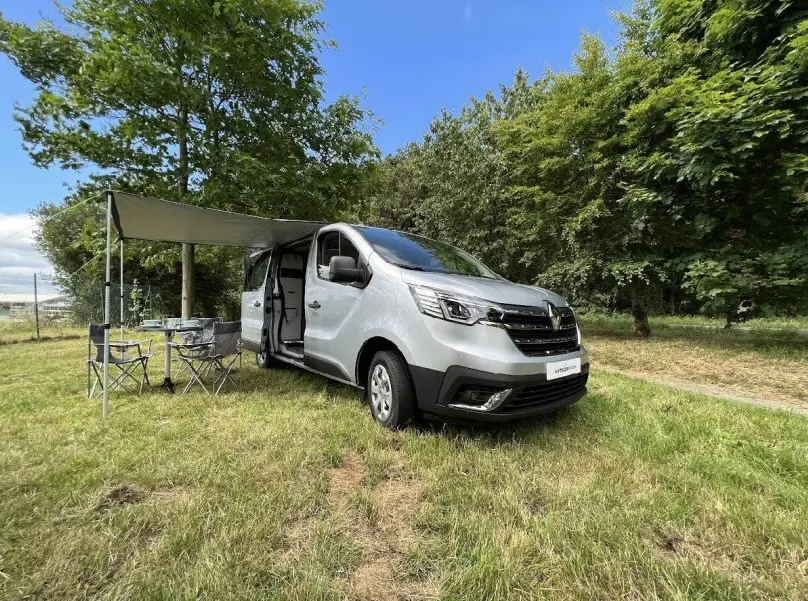 Renault Trafic Fourgon gris en 3/4 avant droit, porte latérale ouverte avec auvent et mobilier de camping installé.
