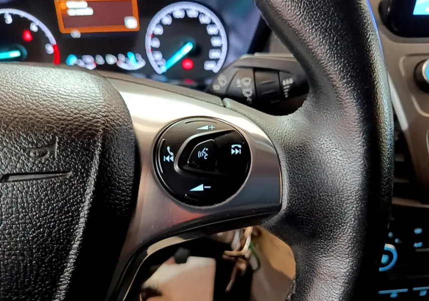 Gros plan sur le volant noir du Ford Transit Fourgon 2022 avec commandes vocales et tableau de bord éclairé en arrière-plan.