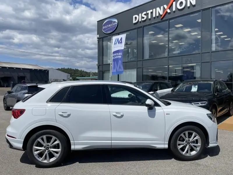Audi Q3 blanc vue de profil côté gauche, finition S-Line avec jantes alliage, stationné devant un concessionnaire.