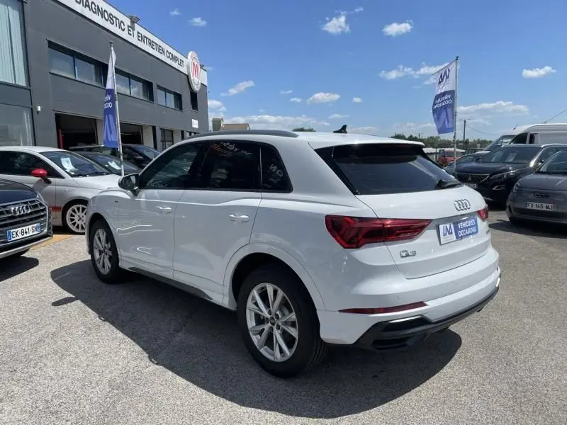 Audi Q3 blanc en vue 3/4 arrière droit, avec vitres teintées et badge S-Line visible sur un parking extérieur.