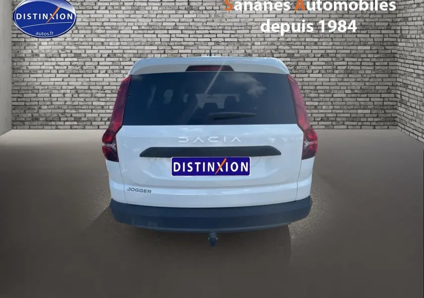 Vue arrière d'un Dacia Jogger blanc avec attelage et vitres arrière surteintées sur fond de mur en briques.