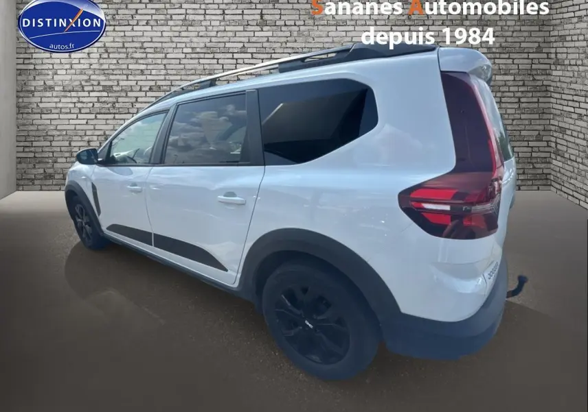 Vue 3/4 arrière droite d'un Dacia Jogger blanc avec jantes noires et attelage fixe sur fond brique.