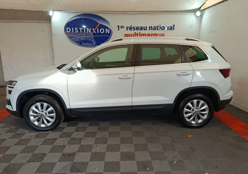 Vue latérale droite d'un Skoda Karoq blanc 2024, avec jantes alliage et barres de toit noires visibles en intérieur showroom.
