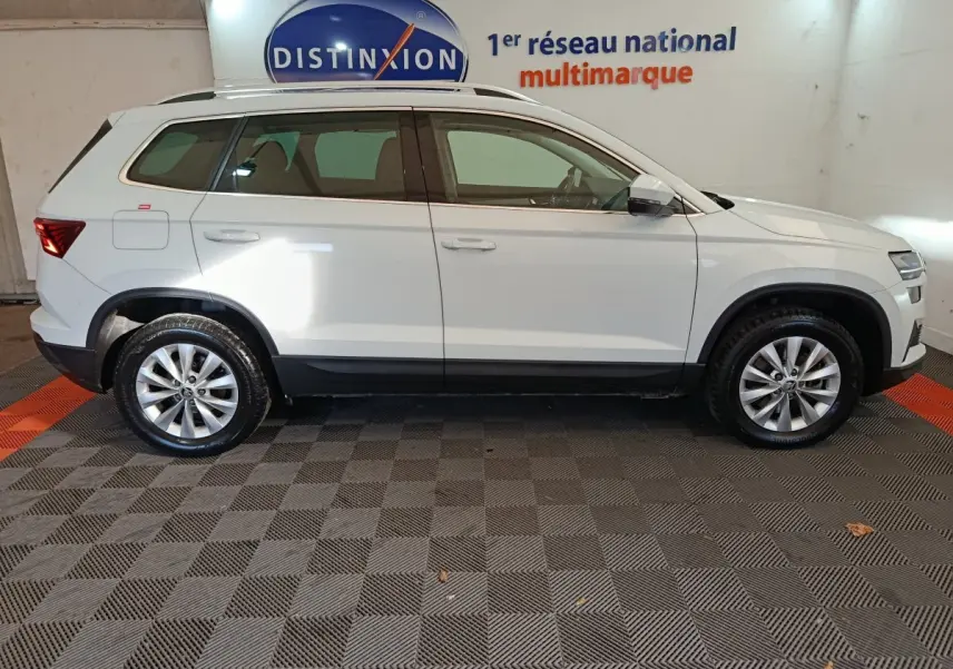 Vue profil droit d'un Skoda Karoq blanc 2024 avec jantes alliage et rails de toit noirs en intérieur showroom.