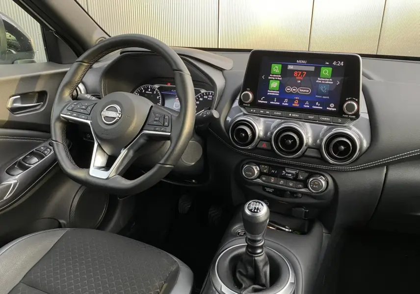 Vue intérieure côté conducteur du Nissan Juke blanc perle 2024 avec volant cuir, écran tactile et boîte manuelle.
