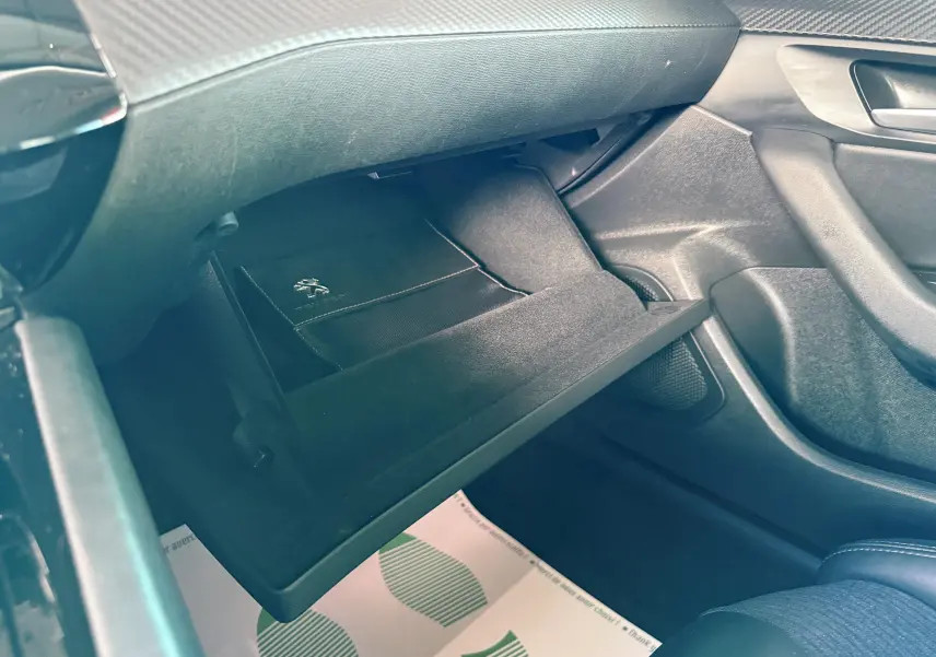 Gros plan sur la boîte à gants ouverte côté passager d'une Peugeot 308 SW noire avec un livret Peugeot à l'intérieur.