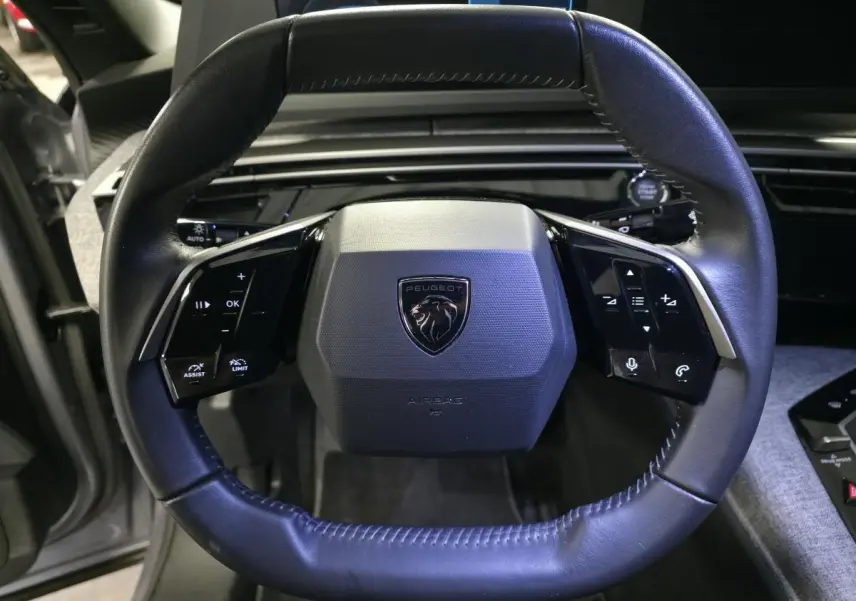 Volant compact en cuir noir avec logo Peugeot au centre, vue frontale rapprochée de l'habitacle gris chiné.