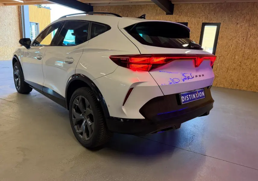 Vue 3/4 arrière droite d'une CUPRA Formentor blanche avec feux arrière allumés dans un garage.