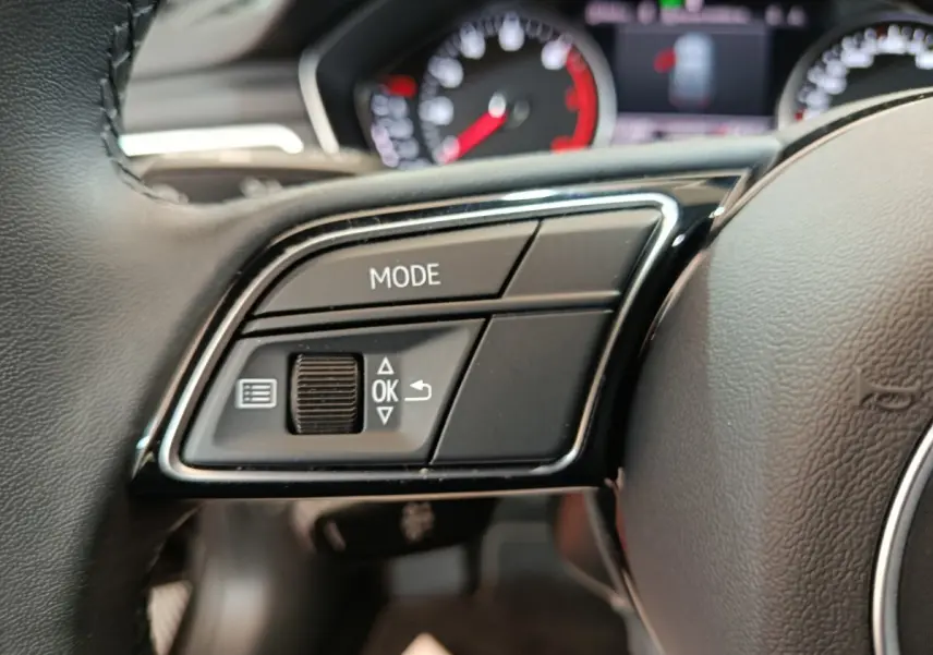 Gros plan sur les commandes gauche du volant cuir noir de l’Audi A4 35 TFSI 2024 avec tableau de bord flou en arrière-plan.