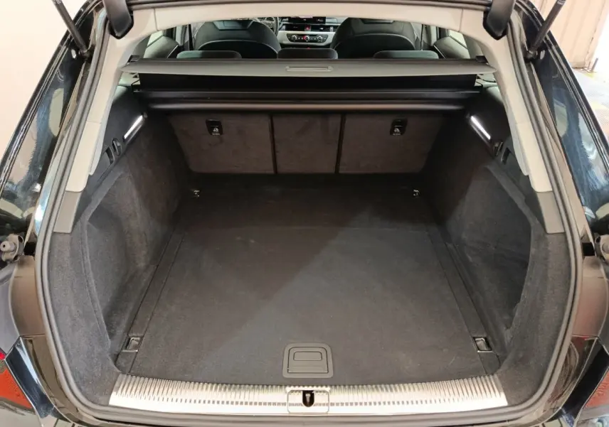 Coffre ouvert vu de l'arrière d'une Audi A4 noire 2024, intérieur spacieux avec tapis noir et éclairage latéral.